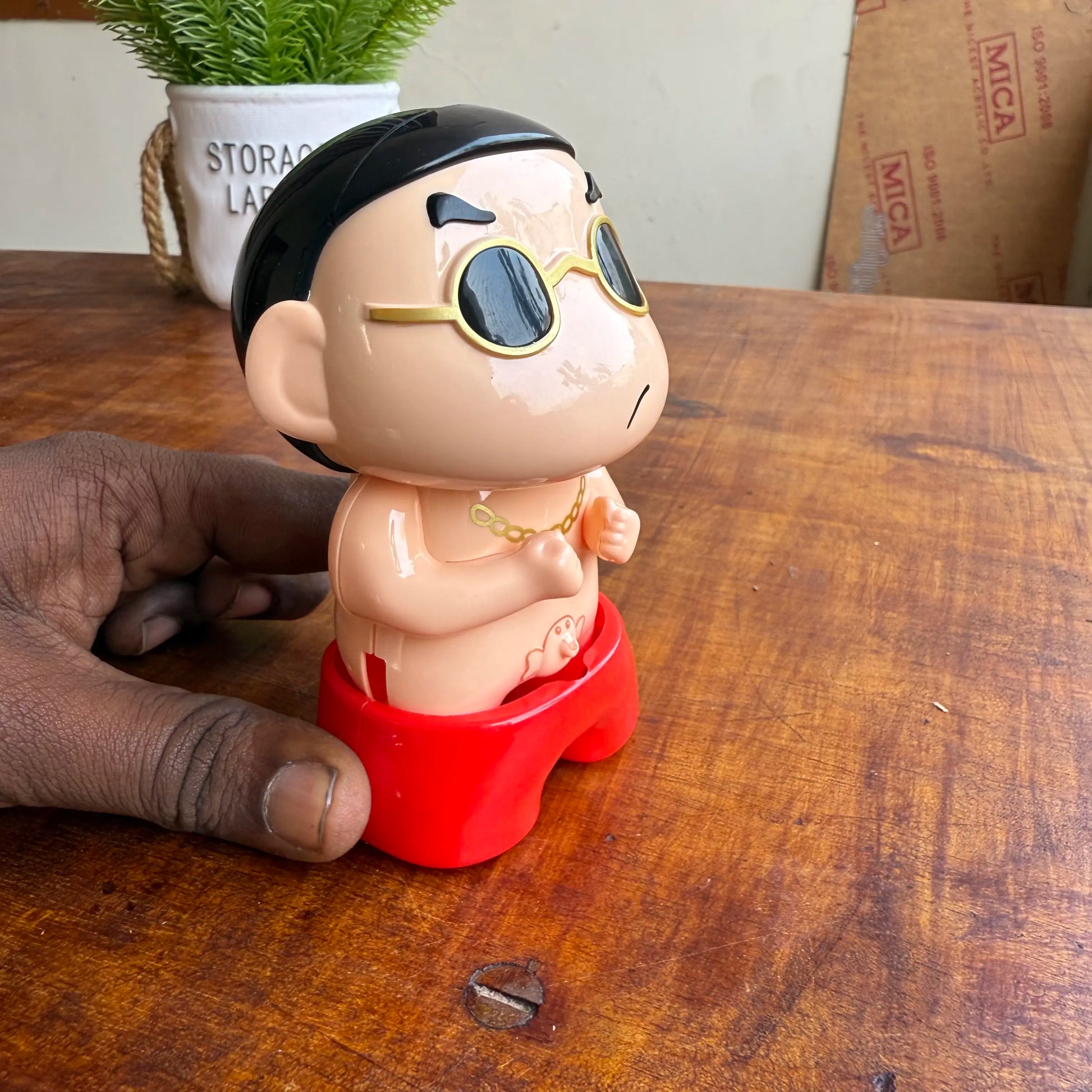 Toy figure of a character shinchan peeing with sunglasses and red base on a wooden surface