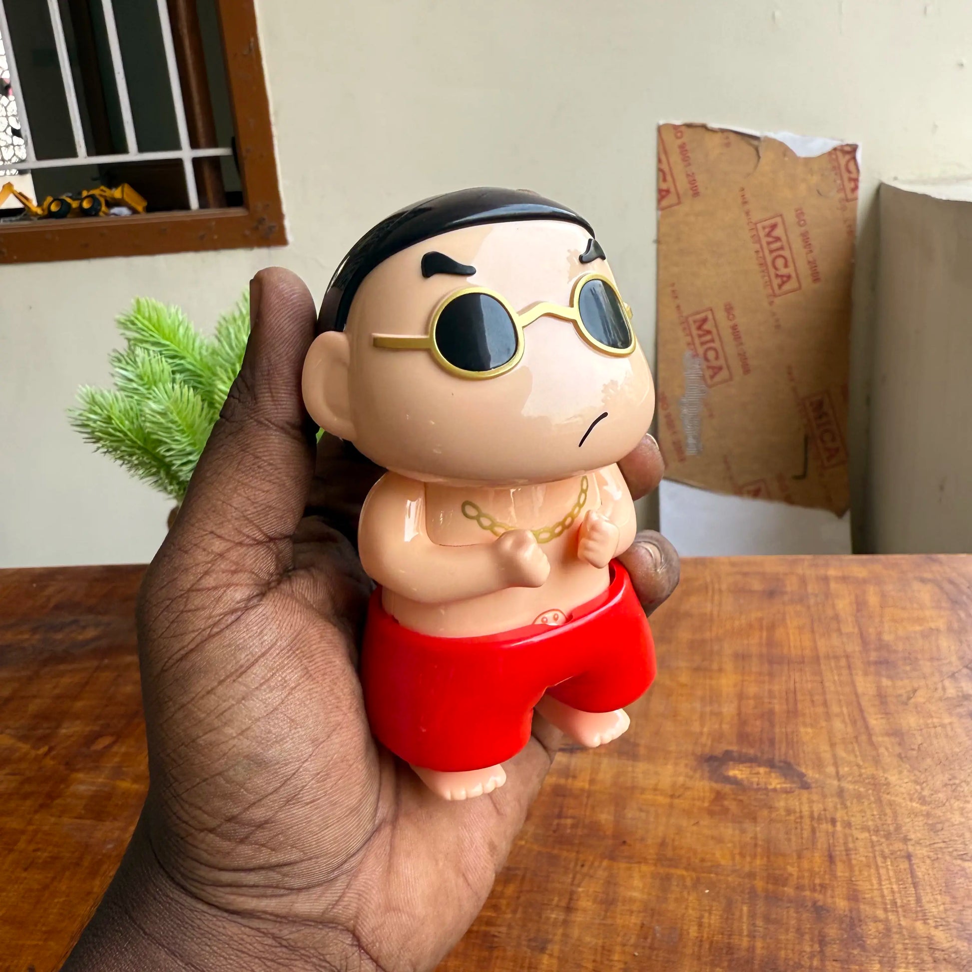 Hand holding a small figurine with sunglasses and red shorts on a wooden surface.
